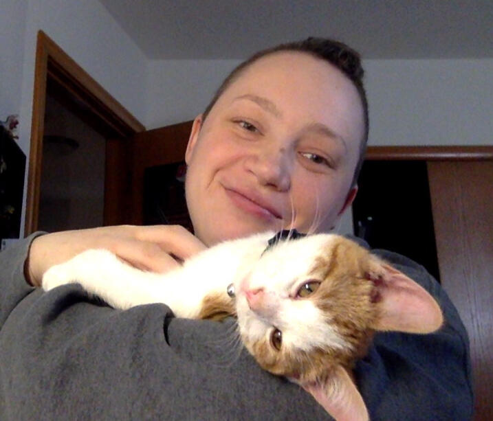 a white non-binary person with short hair is holding a small orange and white kitten with enormous ears, who is draped over their shoulder [Tater Tot as a baby]